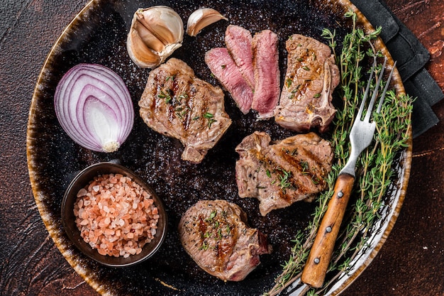 Barbecue Grilled lamb loin chops steaks, saddle in a plate with herbs. Dark background. Top view.