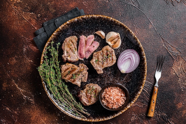 Barbecue Grilled lamb loin chops steaks, saddle in a plate with herbs. Dark background. Top view.