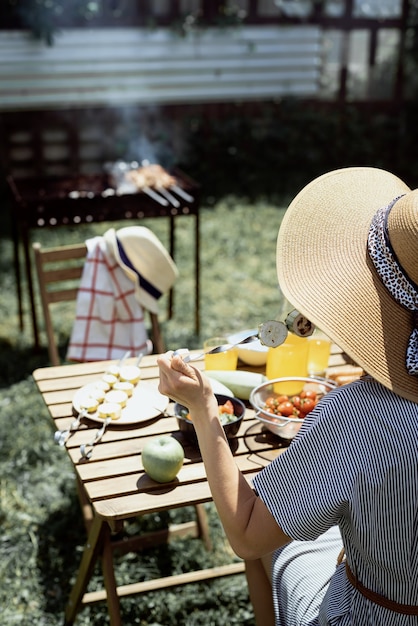 Barbecue in de achtertuin. Jonge vrouw met een zomerhoed die aan tafel zit en buiten gegrilde groenten eet, achteraanzicht