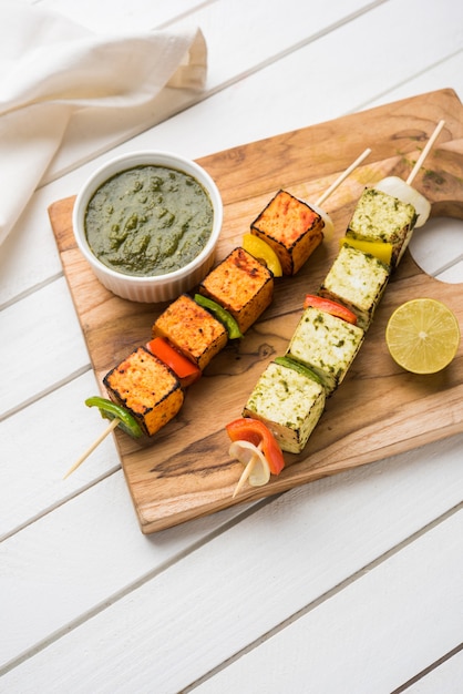 Barbecue Paneer Tikka Kabab - 빨간색과 흰색 소스를 스틱에 넣은 인도 탄두리 코티지 치즈 큐브, 다채로운 또는 나무 테이블 위에 녹색 처트니가 있는 접시에 제공되며 선택적인 초점