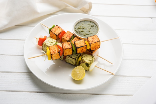 Barbecue Paneer Tikka Kabab - 빨간색과 흰색 소스를 스틱에 넣은 인도 탄두리 코티지 치즈 큐브, 다채로운 또는 나무 테이블 위에 녹색 처트니가 있는 접시에 제공되며 선택적인 초점