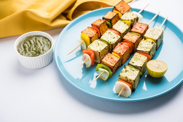 Barbecue Paneer Tikka Kabab - Indian Tandoori cottage cheese cubes in red and white sauce in sticks, served in a plate with green chutney over colourful or wooden table top,selective focus