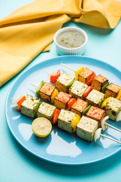 バーベキューパニールティッカカバブ-スティックの赤と白のソースのインドのタンドリーカッテージチーズキューブ、カラフルなまたは木製のテーブルトップの上に緑のチャトニーとプレートで提供、選択的な焦点