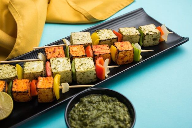 Barbecue Paneer Tikka Kabab - Indian Tandoori cottage cheese cubes in red and white sauce in sticks, served in a plate with green chutney over colourful or wooden table top,selective focus