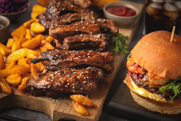 Costine di maiale barbecue su una tavola di legno, spicchi di patate, hamburger e bicchiere di cola, salsa. fast food. avvicinamento.