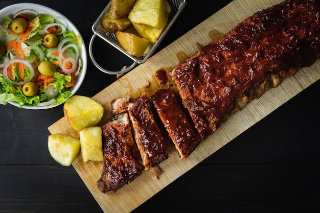 Barbecue ribs with potatoes and salad on a wooden board on a dark background. Copy space.