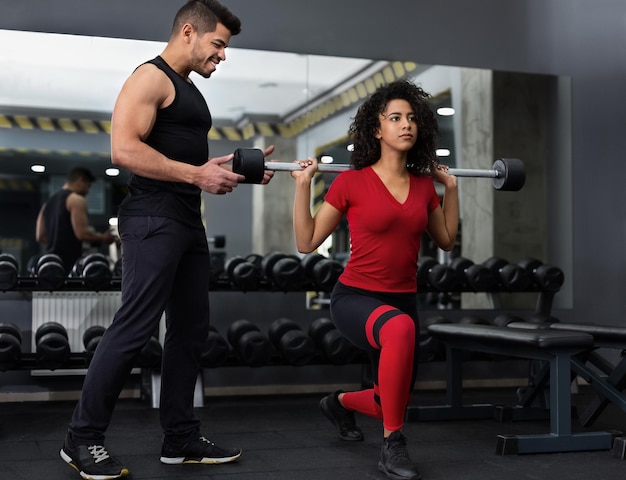 Barbell squats. Personal instructor helping african-american woman in gym