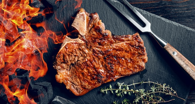 Barbeque steak on a black slate board with meat fork and grill coals