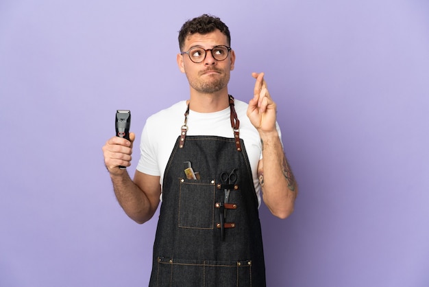 Barber caucasian man in an apron on purple with fingers crossing and wishing the best