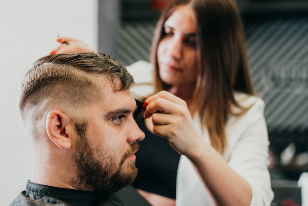 The barber cuts a bearded man with scissors in the salon