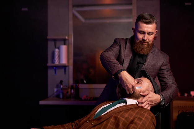 Barber cutting beard of male with razor