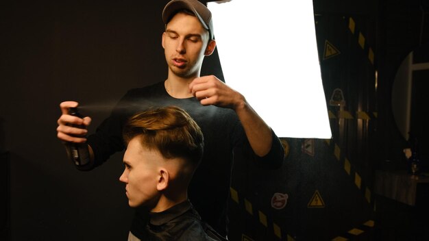 Photo barber makes hair styling with hair spray after haircut at the barber shop young handsome caucasian man getting a haircut in a modern hairsalon from professional hairdresser