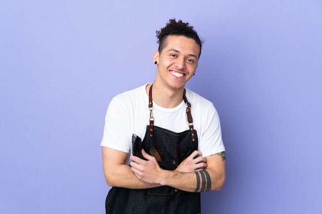 Barber man in an apron over isolated purple background laughing