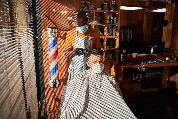 Barber in medical mask cutting client hair in barbershop