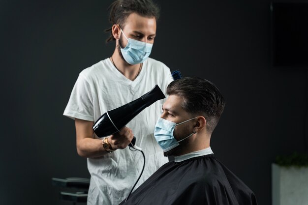 Barber in medical mask dries the hair of a handsome bearded man after a fashionable haircut. The work of a hairdresser during the quarantine period coronavirus covid-19.