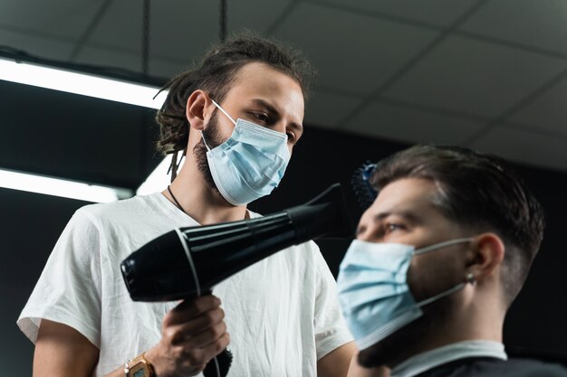 Barber in medical mask dries the hair of a handsome bearded man after a fashionable haircut. The work of a hairdresser during the quarantine period coronavirus covid-19.