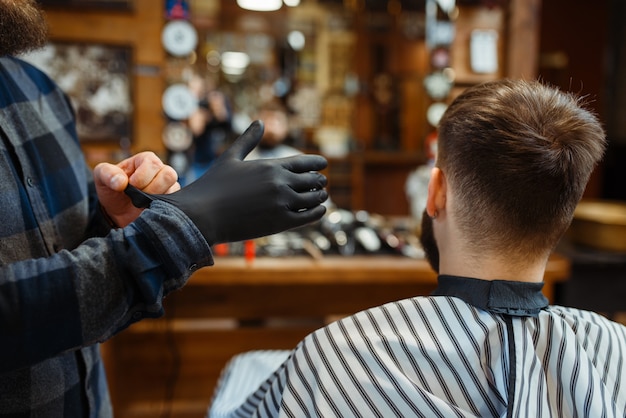 Barber puts on gloves, customer sitting in chair. Professional barbershop is a trendy occupation. Male hairdresser and client in hair salon