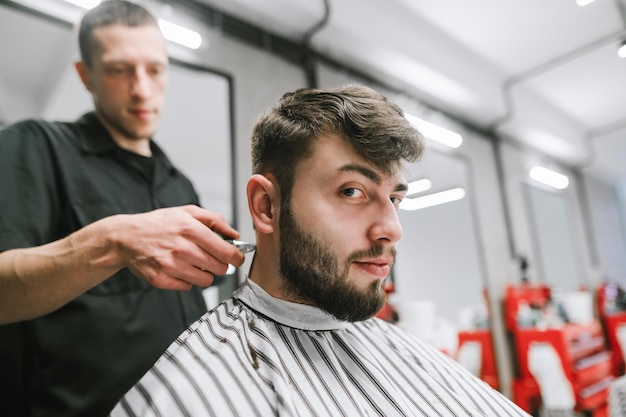 Foto cliente del negozio di barbiere con la barba seduto su una sedia in un peignoir guardando la telecamera