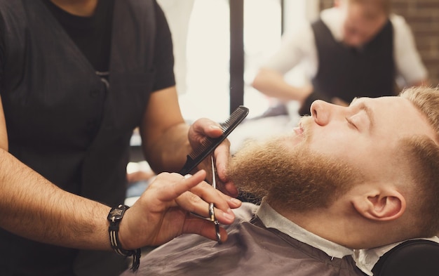 Barber styling beard at barbershop, closeup. Stylish hairdresser in male hair salon cutting hair with scissors