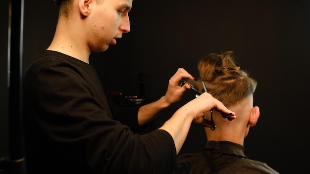 이발사는 현대 이발소에서 젊은 남자 미용사의 갈색 웨이브 머리에 얇은 가위와 금속 빗을 사용합니다.