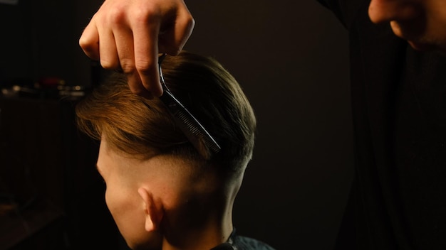 Barber use Thinning scissors and metal comb on brown wavy hair of young man Hairdresser service in a modern barbershop in a dark key lightning with warm light