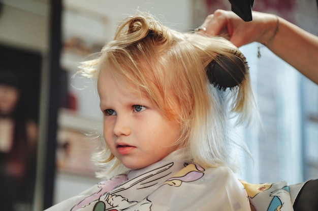 Barber woman make fashionable pretty hairstyle for cute little blond girl child in modern barbershop, hair salon. Hairdresser makes hairdo for young baby in barber shop. Concept hairstyle and beauty