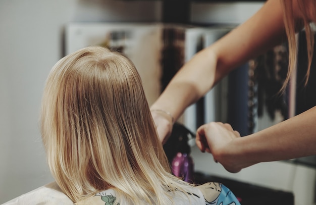 La donna del barbiere fa l'acconciatura graziosa alla moda per la bambina bionda carina nel moderno negozio di barbiere, parrucchiere. il parrucchiere fa la pettinatura per il bambino piccolo nel negozio di barbiere. concept acconciatura e bellezza