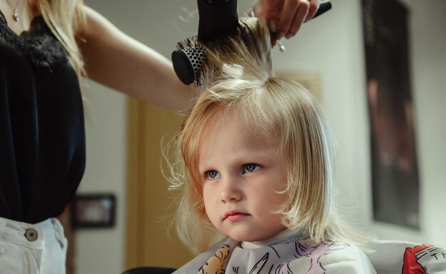 Barber woman make fashionable pretty hairstyle for cute little blond girl