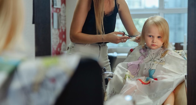 床屋の女性は、かわいい小さなブロンドの女の子のためのファッショナブルなきれいなヘアスタイルを作る