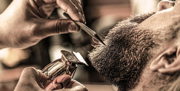 Barber works with a beard clipper. hipster client getting haircut. hands of a hairdresser with a beard clipper, closeup. bearded man in barbershop. man visiting hairstylist in barbershop