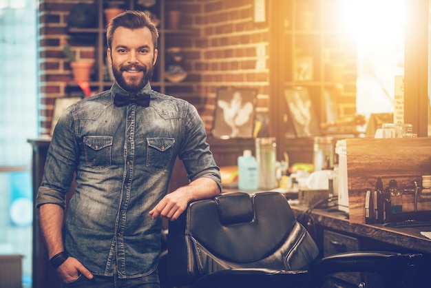 Barbershop owner. Cheerful young bearded man looking at camera and holding hand in pocket while leaning on chair at barbershop