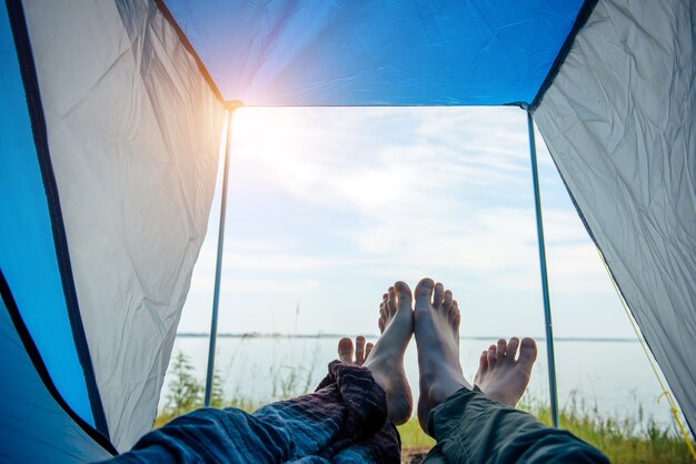 Gambe scoperte dell'uomo e della donna distese fuori dalla tenda turistica. vista sulla sponda del fiume con erba verde e cielo blu in una giornata di sole estivo. attraversato a piedi nudi di amanti che si toccano. turismo familiare, luna di miele.