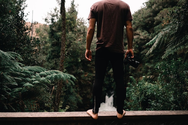 Barefoot blanke jongen in bruin T-shirt en zwarte broek die op de houten reling van een brug staat