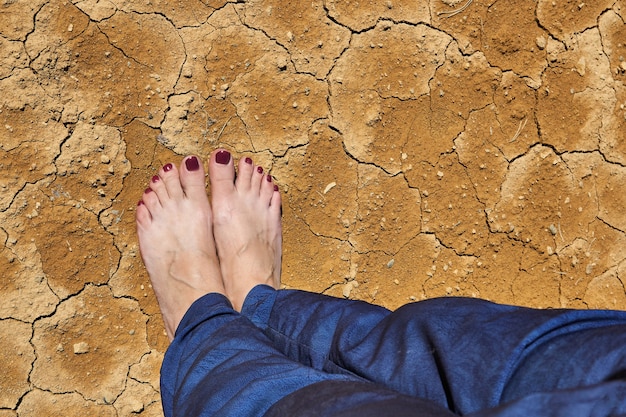 Barefoot female feet in jeans stand on dry cracked clay soil