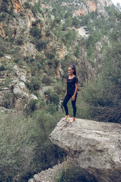 Barefoot female tourist in black clothes standing on stone against mountain range and bushes on summer day in highlands of Mallorca, Spain