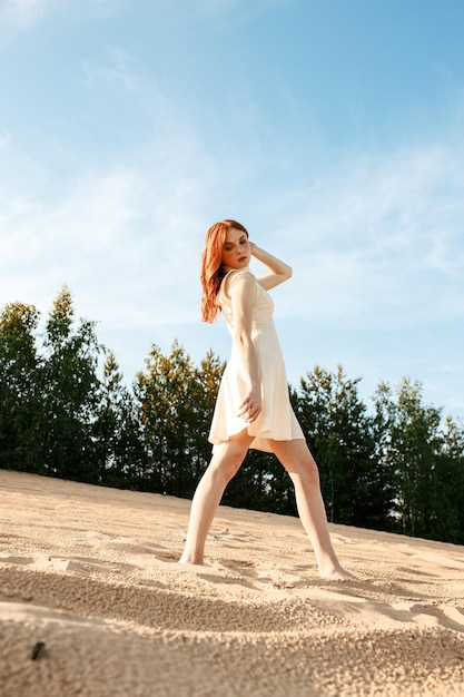 barefoot ginger woman in summer dress standing on sand in nature and touching hair