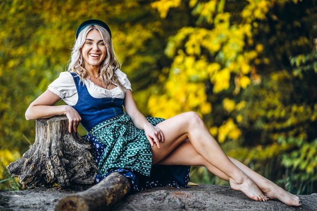 Barefoot happy pretty blond girl in dirndl, traditional beer festival dress, sitting outdoors with blured colorful trees behind