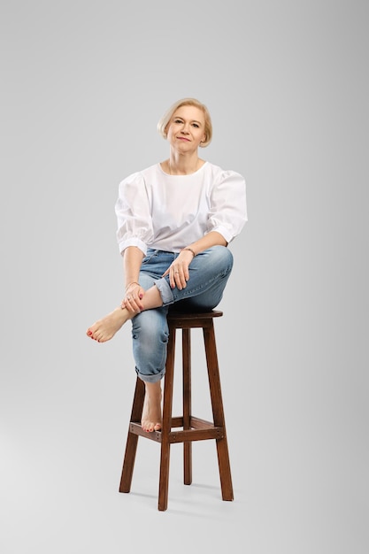 Barefoot senior woman in jeans and white blouse sitting on tall chair in studio throws one leg