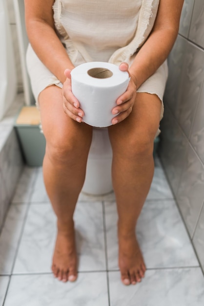 Photo barefoot woman sitting on the toilet with toilet paper