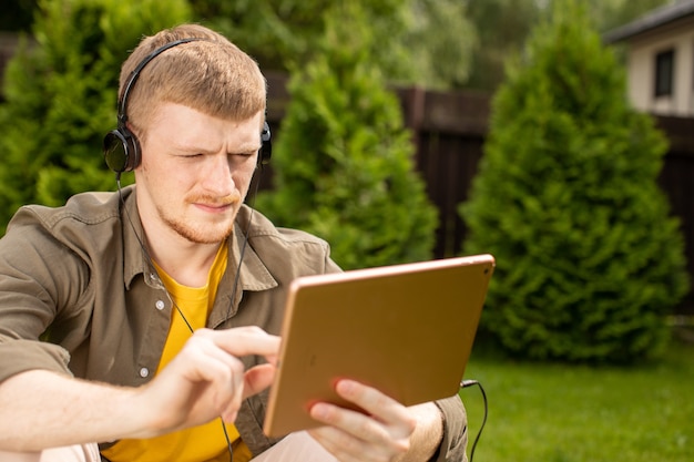 Barefoot young man wearing yellow glasses sitting on green grass with tablet learning foreign languages or listening to music with headphones.