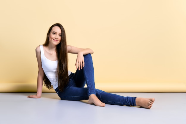 Photo barefoot young woman in white tank top and jeans sitting on the flook in studio