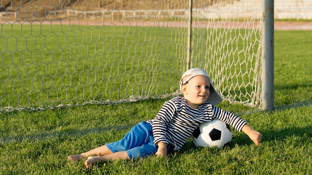 Босоногий мальчик с футбольным мячом, лежащий на зеленой траве перед стойками ворот на спортивном поле