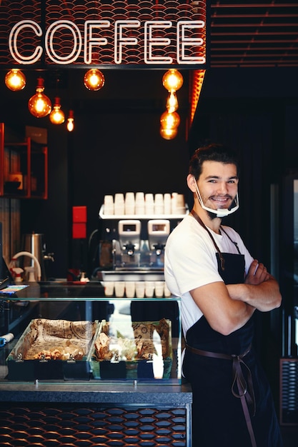 Barista in apron wearing face mask standing at the bar of the coffee shop