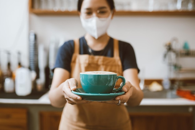 カウンターの後ろに立って、顧客のコーヒーの準備とサービスの概念の中小企業のスタートアップビジネスに提供するためのコーヒーカップを保持しているバリスタアジアの女性