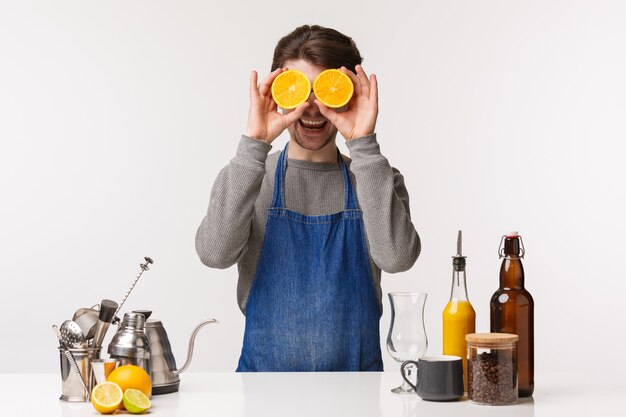 Barista, café werknemer en barman concept. het portret van vrolijke gelukkige mannelijke werknemer die in blauwe schort drank maken, die twee plakken van sinaasappel zoals ogenmasker houden en camera glimlachen, bevindt zich dichtbij barteller