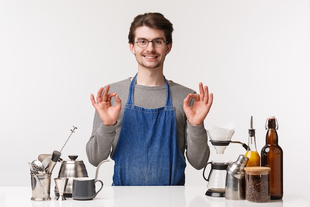 Barista, café werknemer en barman concept. Portret van tevreden, tevreden jonge mannelijke werknemer in schort glimlachend verzekeren, garanderen dat je van koffie houdt, waardoor cappuccino goed teken laat zien