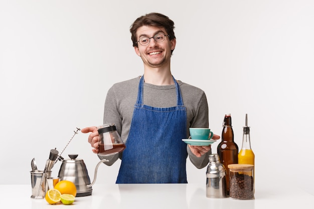Barista, cafe worker and bartender concept. Portrait of cheerful carefree smiling caucasian man in apron, hold kettle with filter coffee and cup, laughing and look away stand bar counter