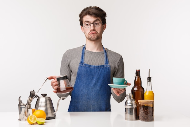 Barista, cafe worker and bartender concept. Portrait of indecisive careless young bored male employee grimacing look away uncertain, hold kettle with filter coffee and cup, 