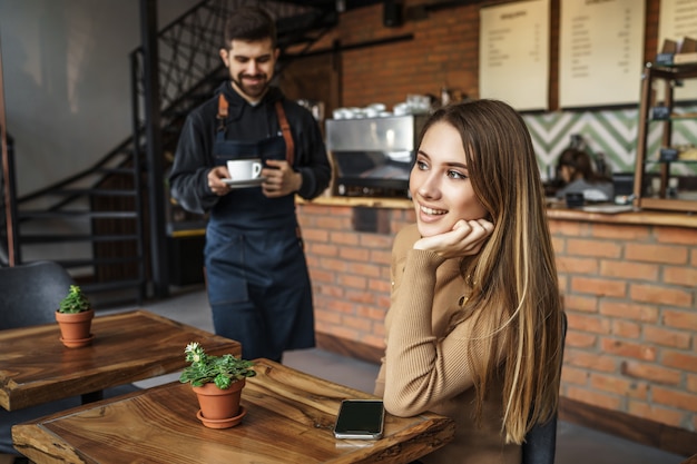 Barista male in blue apron giving order for blonde woman customer in coffee shop