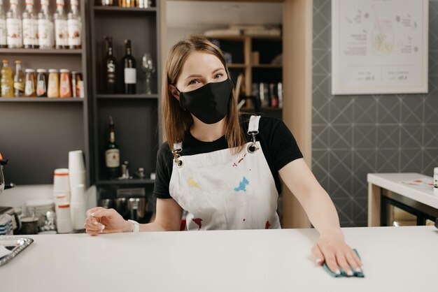 A barista in a medical black face mask smiles and cleaning the bar counter in a coffee shop